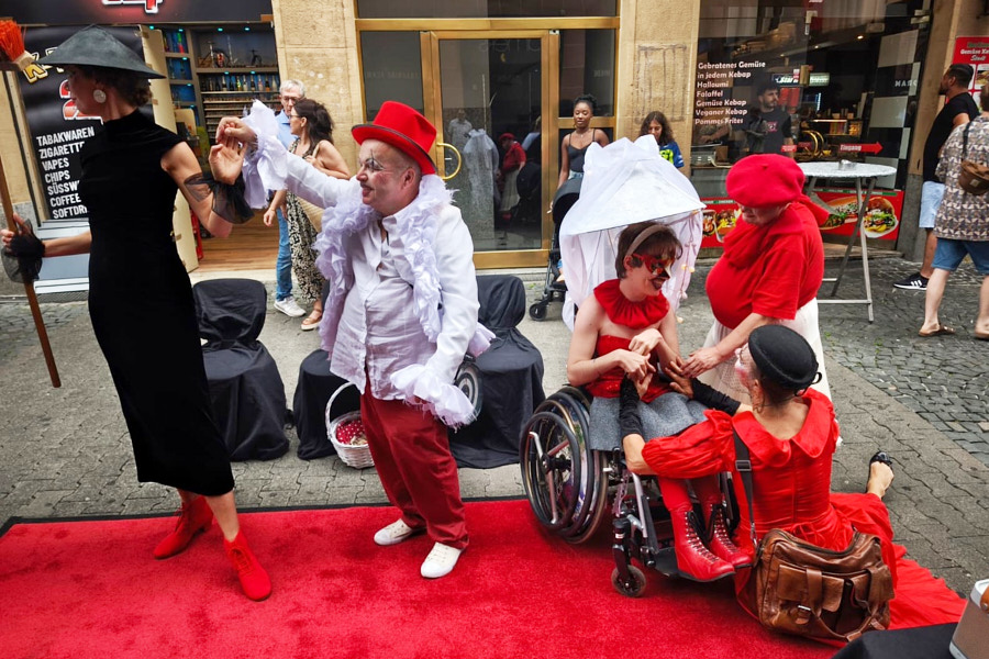 Eine Gruppe von Performance-Künstlern in rot-weißen und schwarzen Kostümen agiert auf einem roten Teppich in einer Fußgängerzone. Die Szene wirkt wie ein lebendiges Theaterstück, bei dem eine Person im Rollstuhl in die Interaktion mit den anderen maskierten und geschminkten Darstellern eingebunden ist.