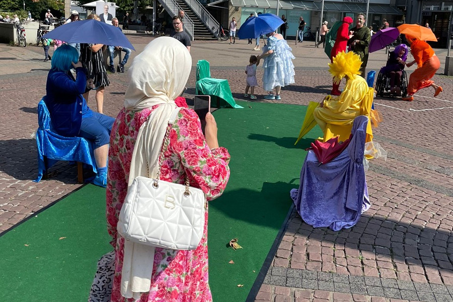 Eine Frau im Vordergrund fotografiert mit ihrem Handy eine Performance auf einem öffentlichen Platz. Entlang eines schmalen grünen Teppichs sitzen und stehen verschiedene Darsteller in auffälligen, einfarbigen Kostümen (Blau, Gelb, Rot, Weiß), die farblich passende Schirme und Tücher verwenden.