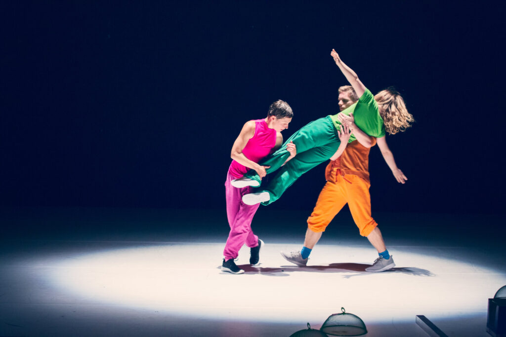 Drei Tänzer in leuchtend pinker, grüner und oranger Kleidung führen eine Hebefigur auf einer dunkel beleuchteten Bühne aus. Zwei Personen halten eine dritte Person in der Waagerechten, während ein heller Spot die Szene von unten beleuchtet.