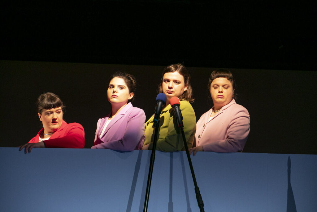 Vier Darstellerinnen stehen nebeneinander hinter einer hohen, hellblauen Brüstung auf einer Theaterbühne. Sie tragen formelle Blazer in kräftigen Farben (Rot, Flieder, Hellgrün und Rosa). Vor ihnen stehen zwei Mikrofone auf Stativen, während sie mit ernsten Mienen ins Publikum blicken.