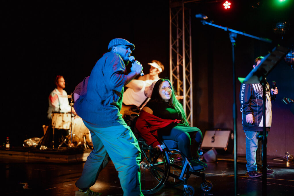 Eine lebendige Live-Performance auf einer dunklen Bühne. Im Vordergrund rockt ein bärtiger Sänger mit einer grauen Schiebermütze, einer weiten blauen Jacke und einer beigefarbenen Hose ins Mikrofon. Neben ihm, in der Mitte, strahlt eine Frau im Rollstuhl. Sie trägt eine schwarze Jacke über einem roten Oberteil und eine rote Gesichtsmaske am Hals. Im Hintergrund sind weitere Bandmitglieder zu sehen: ein Schlagzeuger links, ein Gitarrist in der Mitte und eine weitere Person mit einem Mikrofon rechts. Die Bühne ist von bunten Scheinwerfern in Blau, Rot und Grün beleuchtet, die eine energiegeladene Atmosphäre erzeugen.