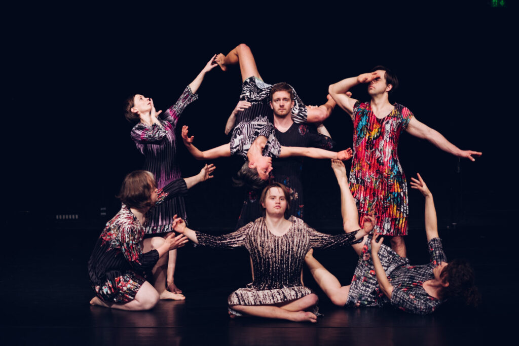 Ein Ensemble-Porträt von sieben Tänzerinnen und Tänzern in einer komplexen, skulpturalen Formation auf einer schwarzen Bühne. Alle tragen unterschiedliche, bunt gemusterte oder strukturiert gestreifte Kleider und sind barfuß. Im Zentrum sitzt eine Person mit Down-Syndrom im Schneidersitz, die Arme weit ausgebreitet. Über ihr wird eine Tänzerin kopfüber in der Luft gehalten. Die anderen Gruppenmitglieder knien oder stehen drumherum, wobei ihre Arme und Blicke in verschiedene Richtungen weisen, was eine harmonische und kraftvolle Gesamtkomposition erzeugt.