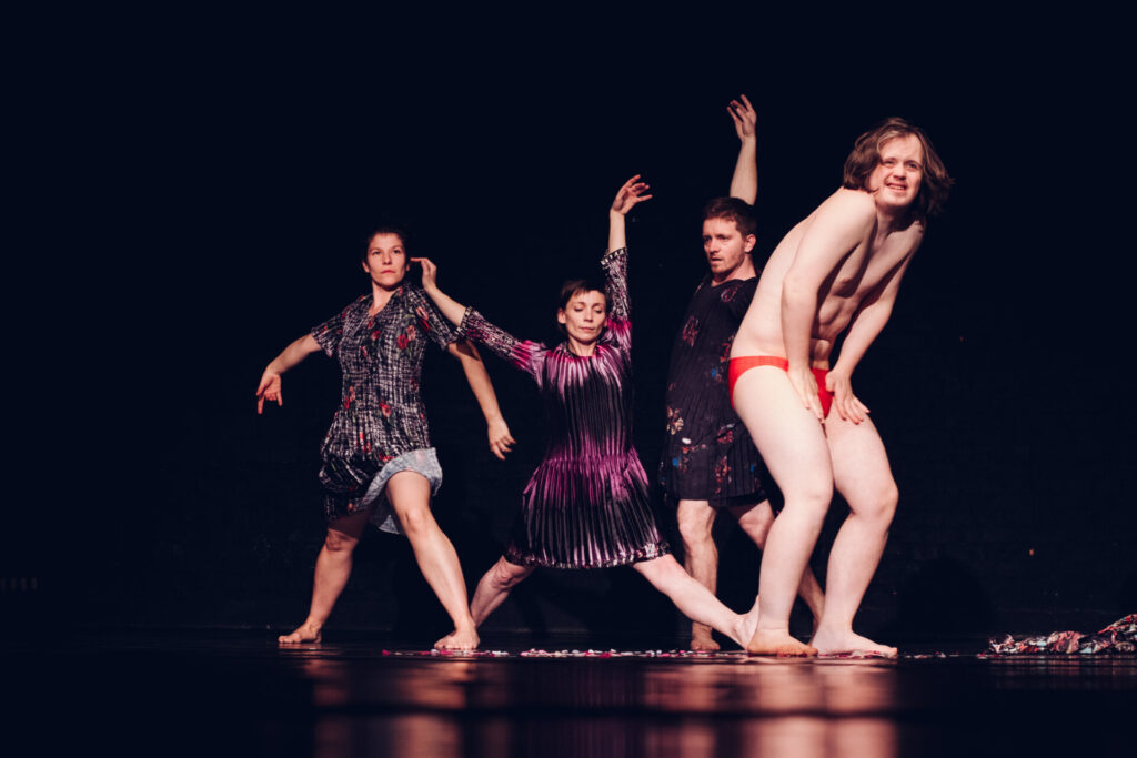 Vier Tänzer:innen stehen auf einer dunklen Bühne in einer choreografierten Pose. Drei tragen kurze, schimmernde Kleider, während eine Person rechts nur rote Unterwäsche trägt. Die Gruppe bewegt sich synchron mit ausgestreckten Armen und gebeugten Knien. Der Bühnenboden ist leicht reflektierend und mit verstreuten Stoffstücken bedeckt, der Hintergrund bleibt schwarz und lenkt den Fokus auf die Körper und Bewegungen.